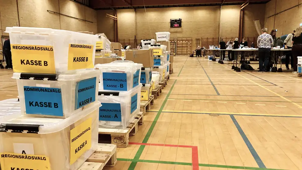 Billede af kasser med stemmer i en sportshal, med stemmetællere i baggrunden