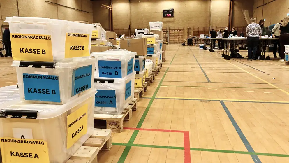 Billede af kasser med stemmer i en idrætshal med mennesker, der tæller stemmer i baggrunden