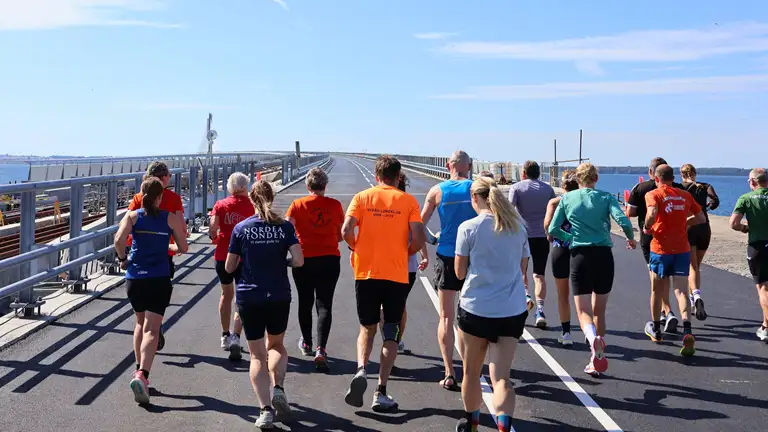 En gruppe mennesker iført sportstøj løber over en bro i solskinsvejr.